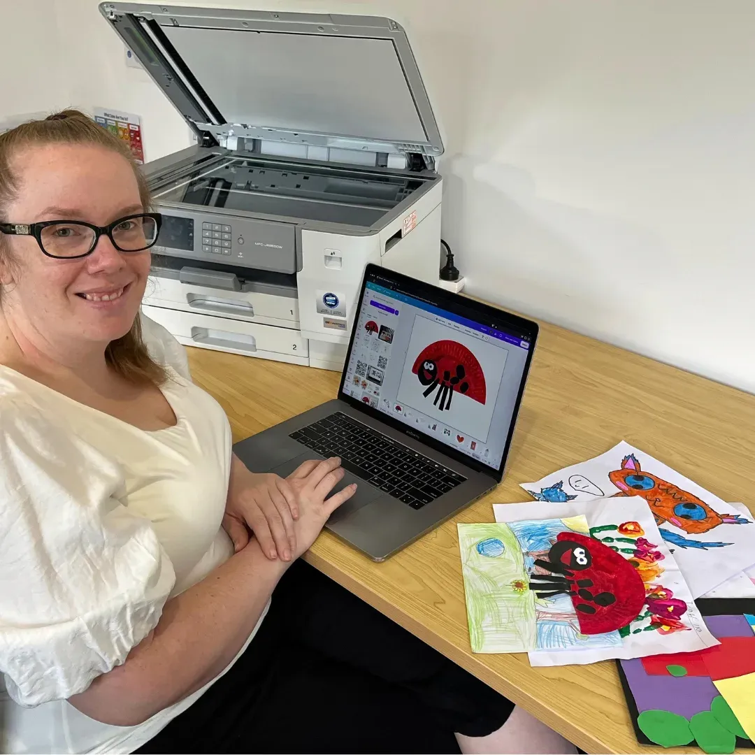 Tamara smiling at her desk, a laptop showing a child's ladybug artwork, an open scanner behind her and children's drawings spread beside her